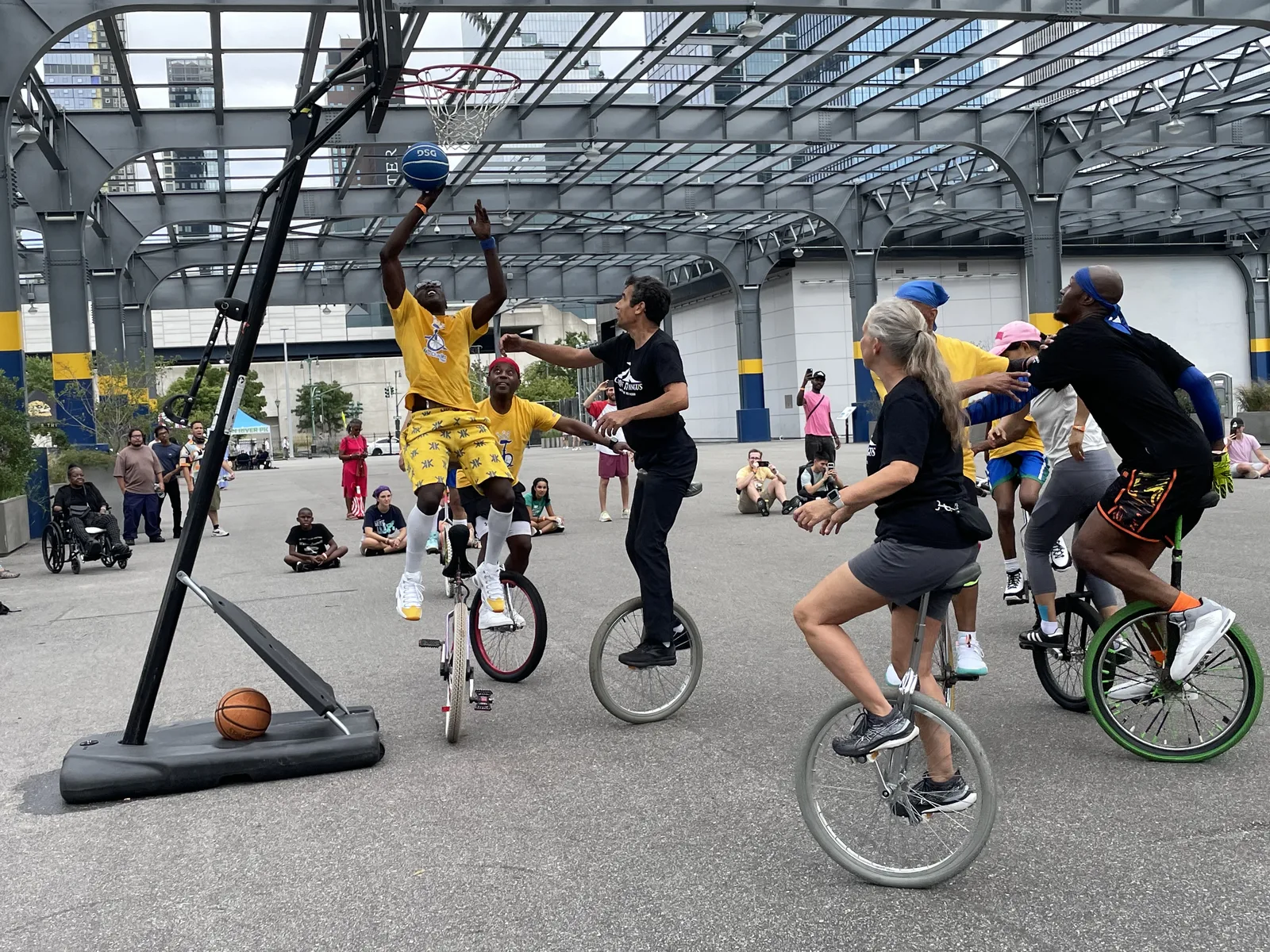 Unicyclists contest a basketball play beneath an outdoor hoop at Pier 76