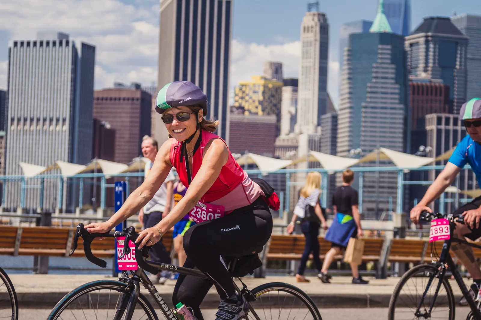 A cyclist in a red jersey rides with the Lower Manhattan skyline behind her