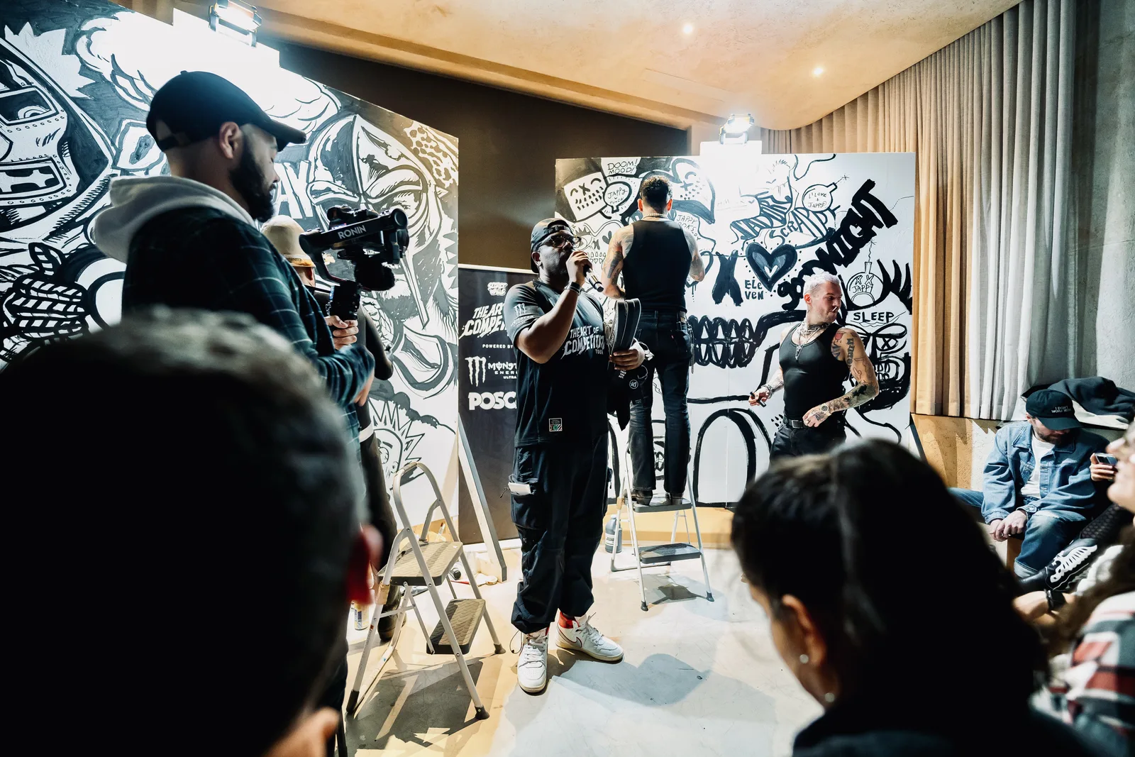 A host addresses the crowd while artists work on black-and-white murals behind him