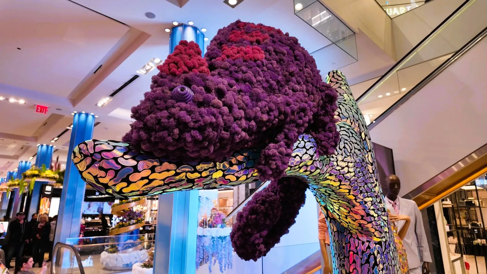 A large purple flower-covered animal sculpture rests on an iridescent branch inside Macy's