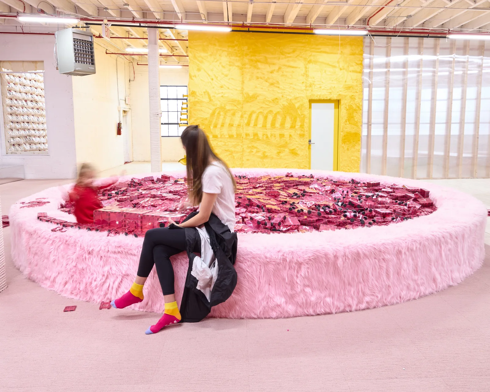 A visitor and a child beside a circular pink play pit filled with transparent building pieces at Space Club DUMBO