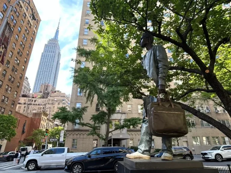 A fragmented bronze traveler carrying a suitcase stands on Park Avenue with the Empire State Building behind it