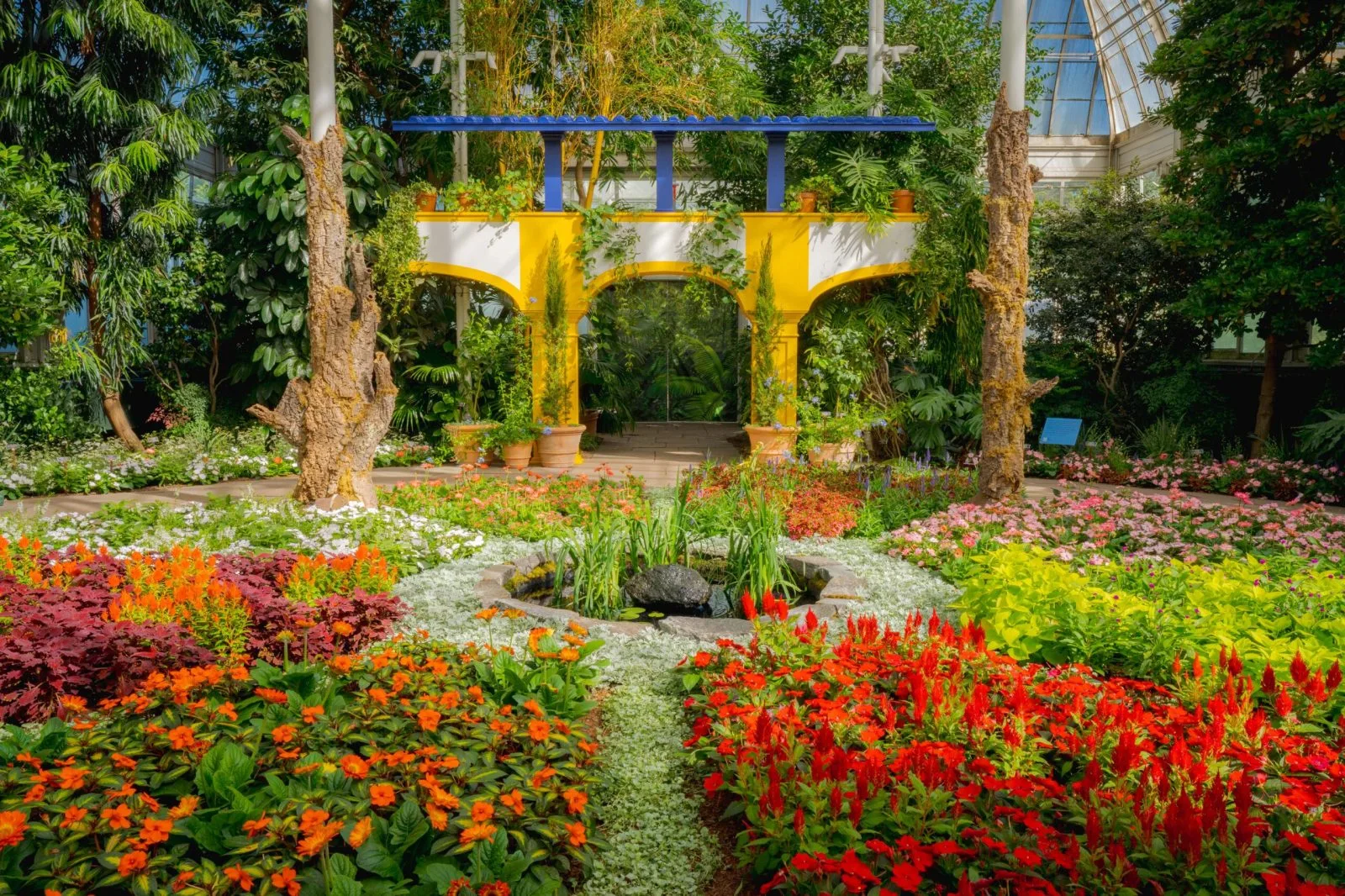 Red, orange, white, and pink flowers surround a yellow garden structure inside the conservatory