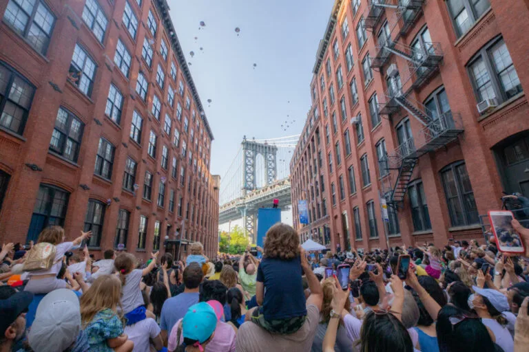 A crowd watches toy elephants descend between brick buildings on Washington Street with the Manhattan Bridge behind them