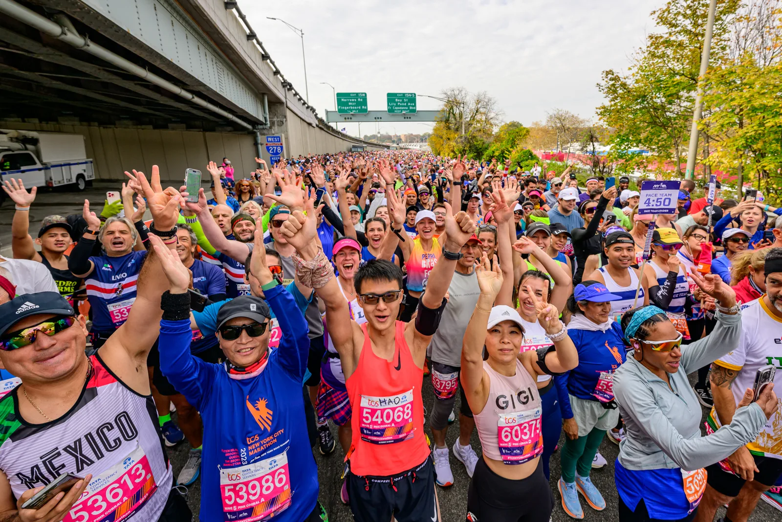 A dense field of New York City Marathon runners raises their hands while moving through Brooklyn
