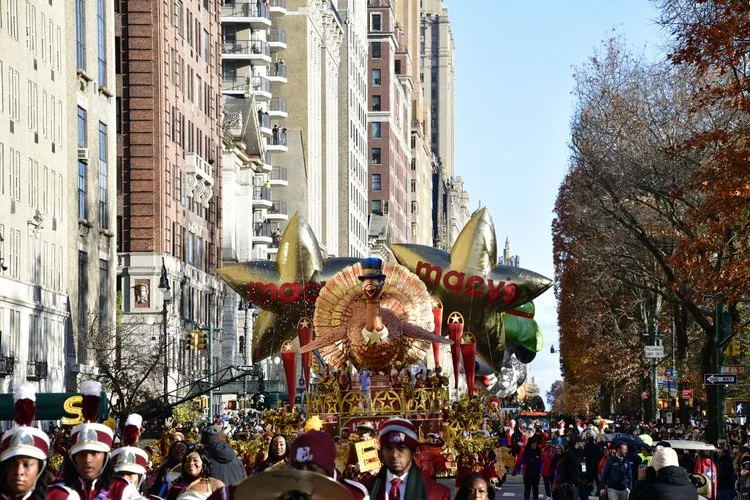 The Tom Turkey float moving down Central Park West with parade balloons, performers, and spectators