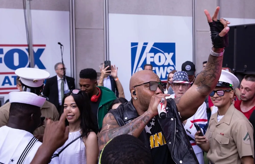 Flo Rida sings into a microphone while raising one arm amid a crowd at FOX Square