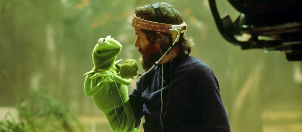 Against a green, forest background, a man with a beard, bandana, and microphone holds a green felt frog puppet in his arm and they man and muppet look at each other fondly.