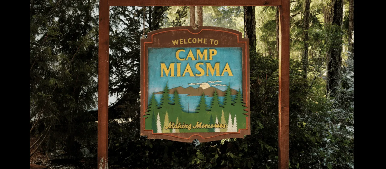 A rustic wooden sign hangs from a wooden frame in a forest setting. The sign reads "Welcome to Camp Miasma. Making Memories" over an image of pine trees, mountains, and a lake.