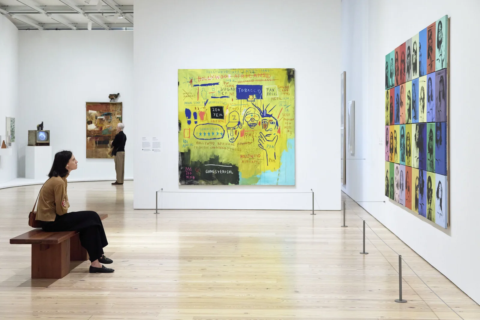 A woman sits on a bench looking at large colorful paintings in a modern art gallery.